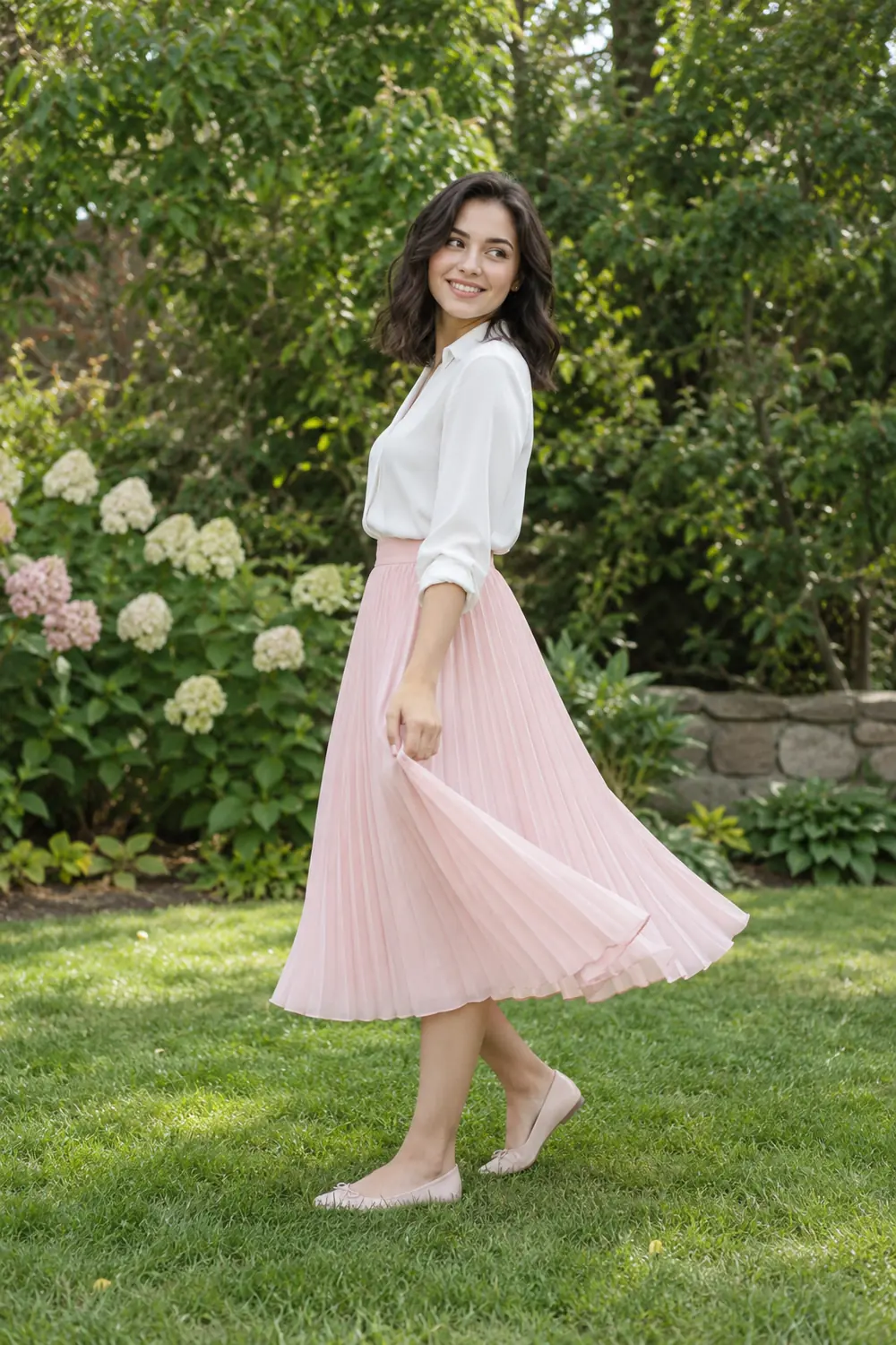 A smiling adult woman with shoulder-length dark hair gently turns in a pale-pink pleated skirt, white blouse, and flats on a green garden lawn.