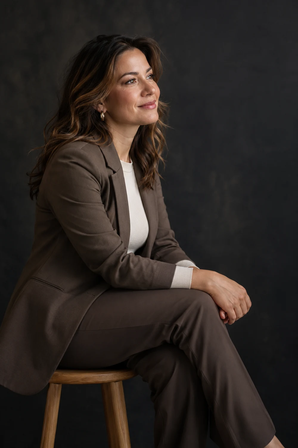 A woman in a brown blazer and trousers sits on a wooden stool against a dark studio backdrop, looking toward soft light.