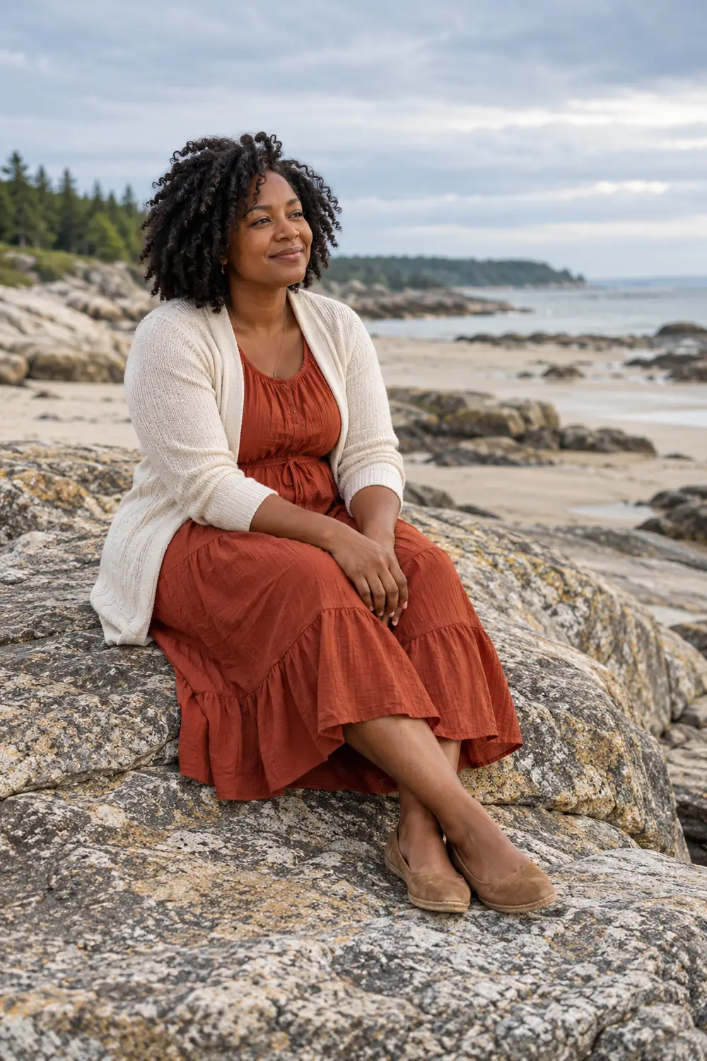 Adult woman with natural coily hair seated on a broad shoreline rock in a rust midi dress, light cardigan, and closed flats.