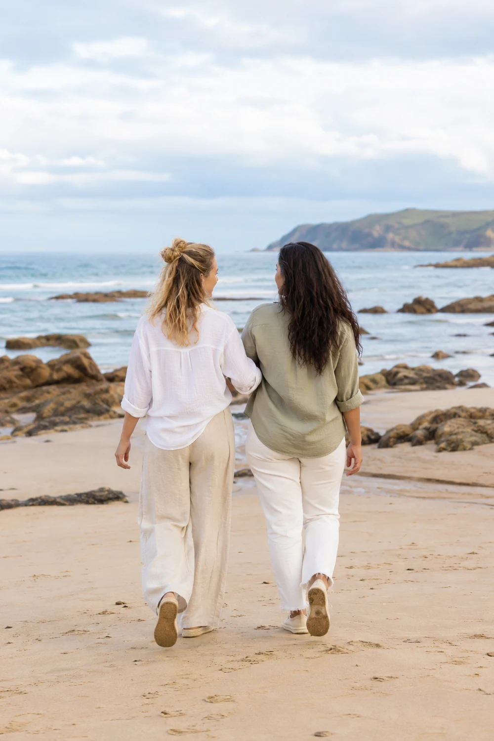Two adult women in light shirts and pale trousers walking arm in arm across the sand toward a rocky sea, seen from behind.