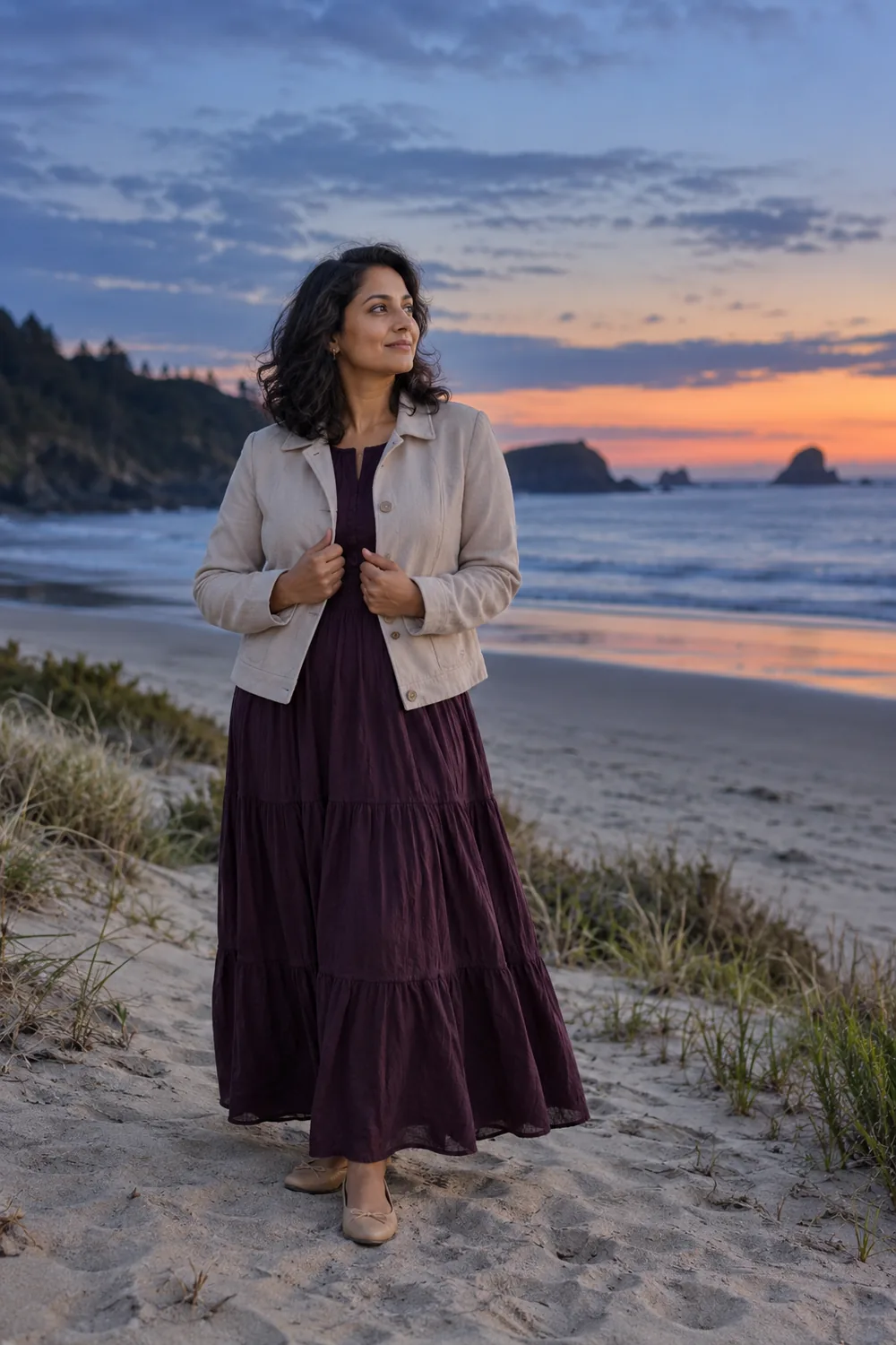 A woman in a plum dress and light jacket stands on sandy beach at twilight, looking toward a calm sea and distant rocky shoreline.