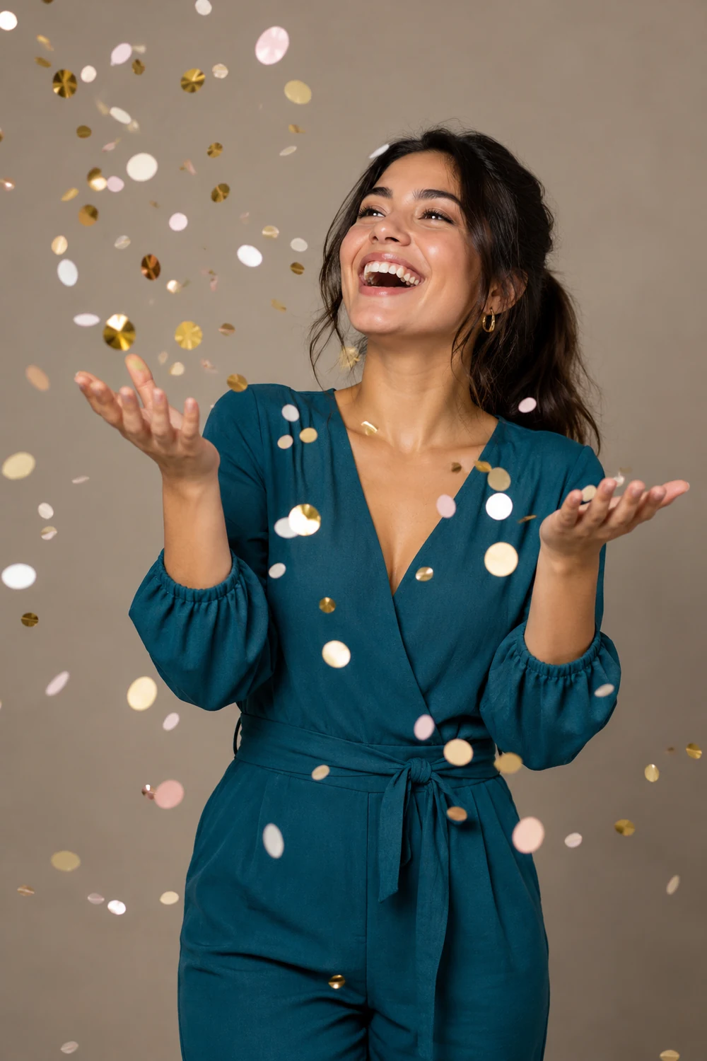 A woman in a teal jumpsuit laughs as she tosses gold, pink, and white confetti in a warm-gray studio.