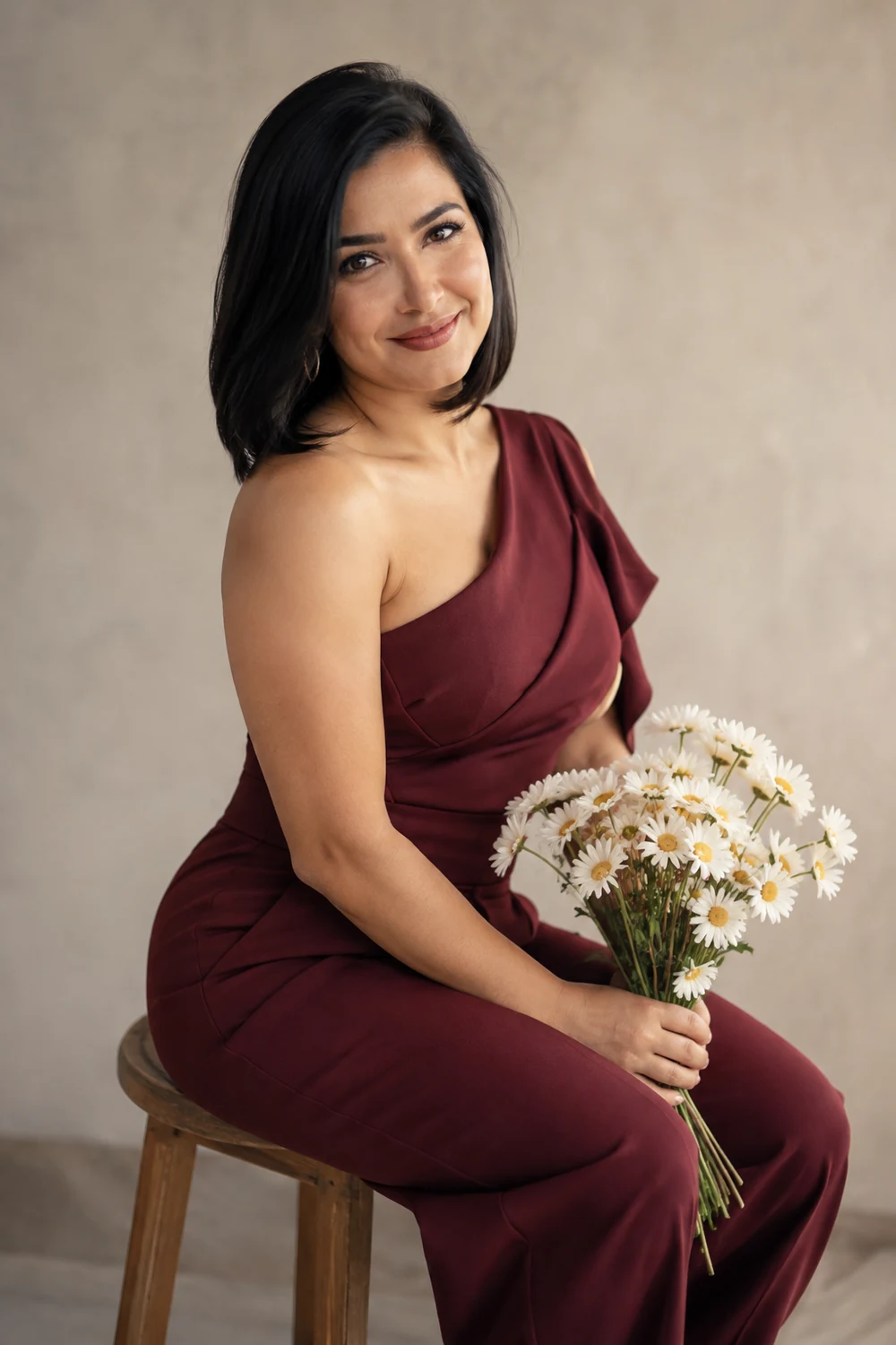 A woman with a black bob sits on a stool in a burgundy one-shoulder jumpsuit, turning toward the camera with daisies in her lap against a neutral studio background.