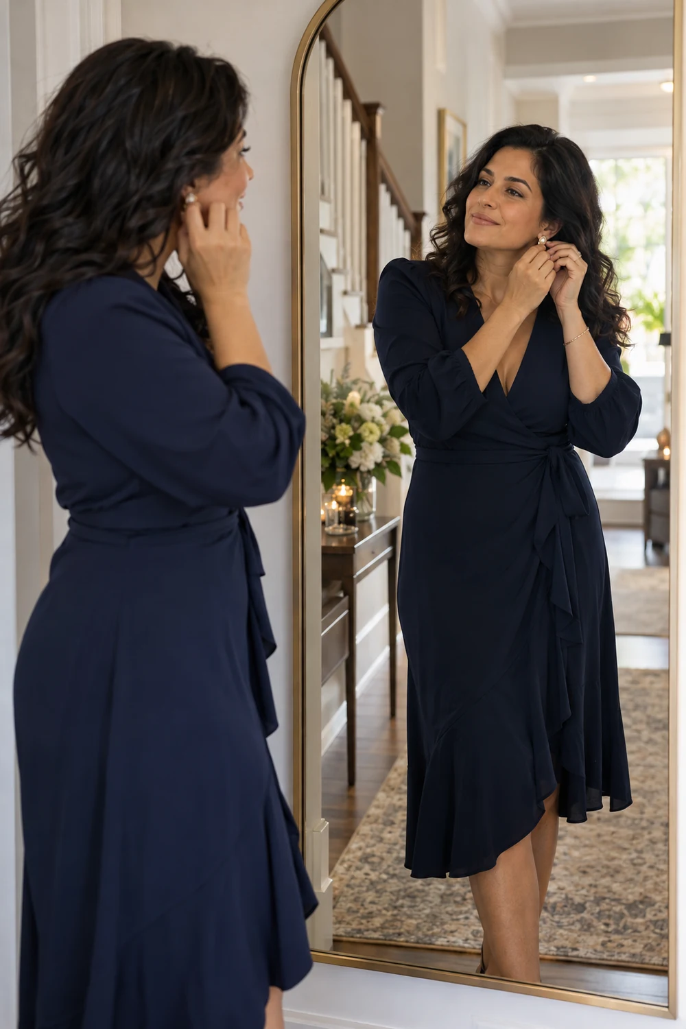 Woman in a navy wrap dress adjusting an earring beside her reflection in a full-length hallway mirror.
