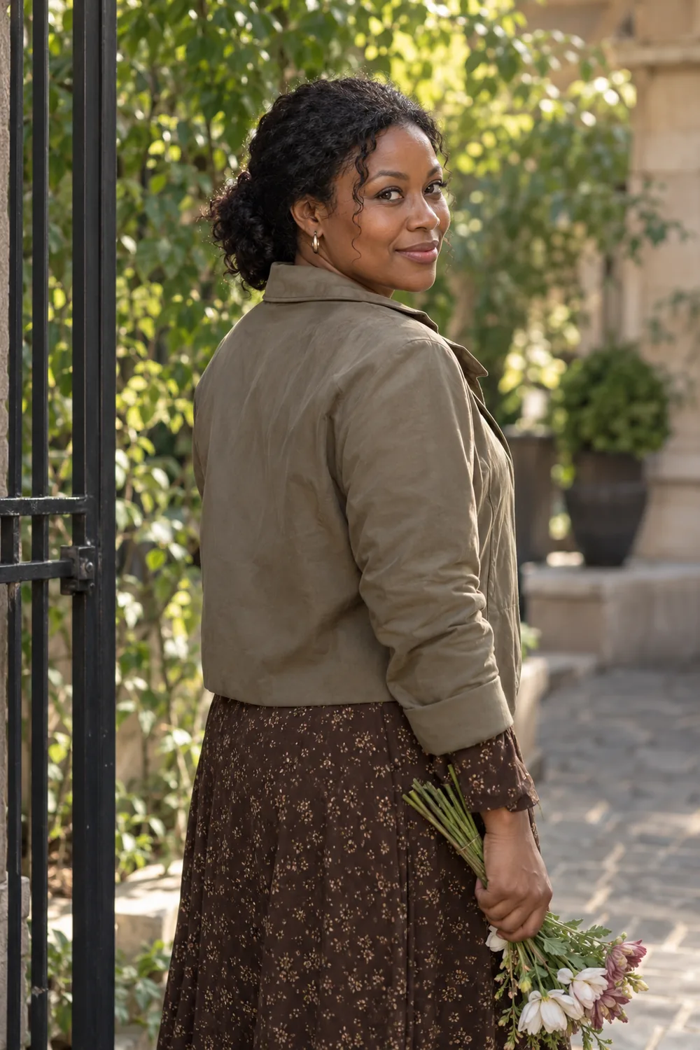 Adult woman in a light jacket and patterned dress turning over her shoulder beside a courtyard gate, holding flowers at her side.