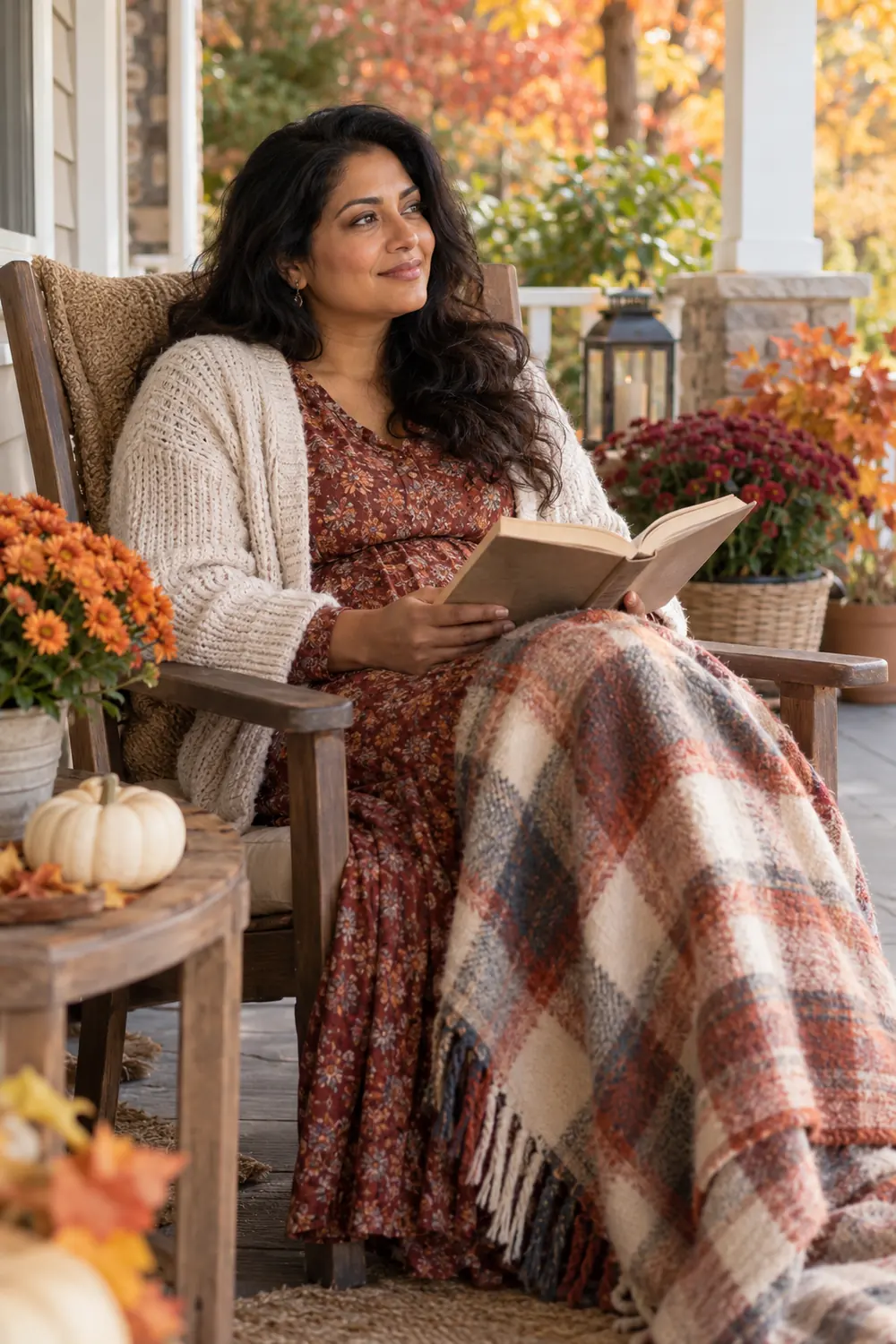 A woman in a cardigan sits on a covered porch with an open book and knitted throw, surrounded by autumn flowers and potted foliage.