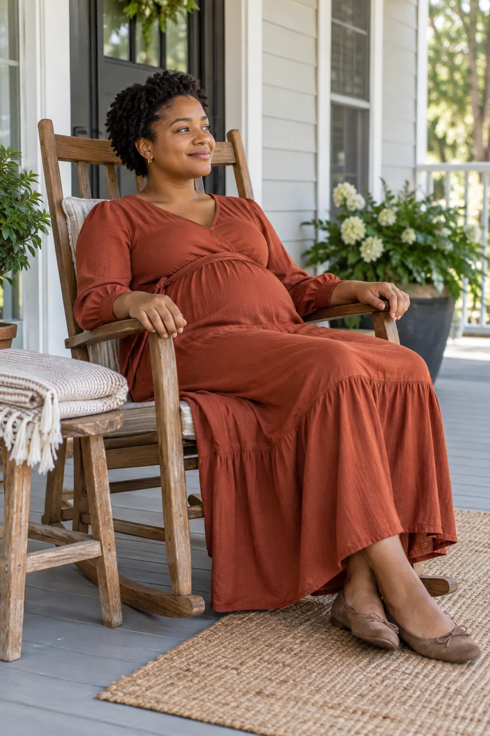 A pregnant woman with short coiled hair sits in a wooden rocking chair on a porch, wearing a rust-colored maternity dress, with folded neutral blankets nearby.