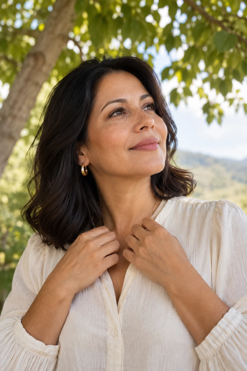 A woman in a cream blouse looks calmly toward the open sky beneath a leafy tree, with her hands resting near her collar.