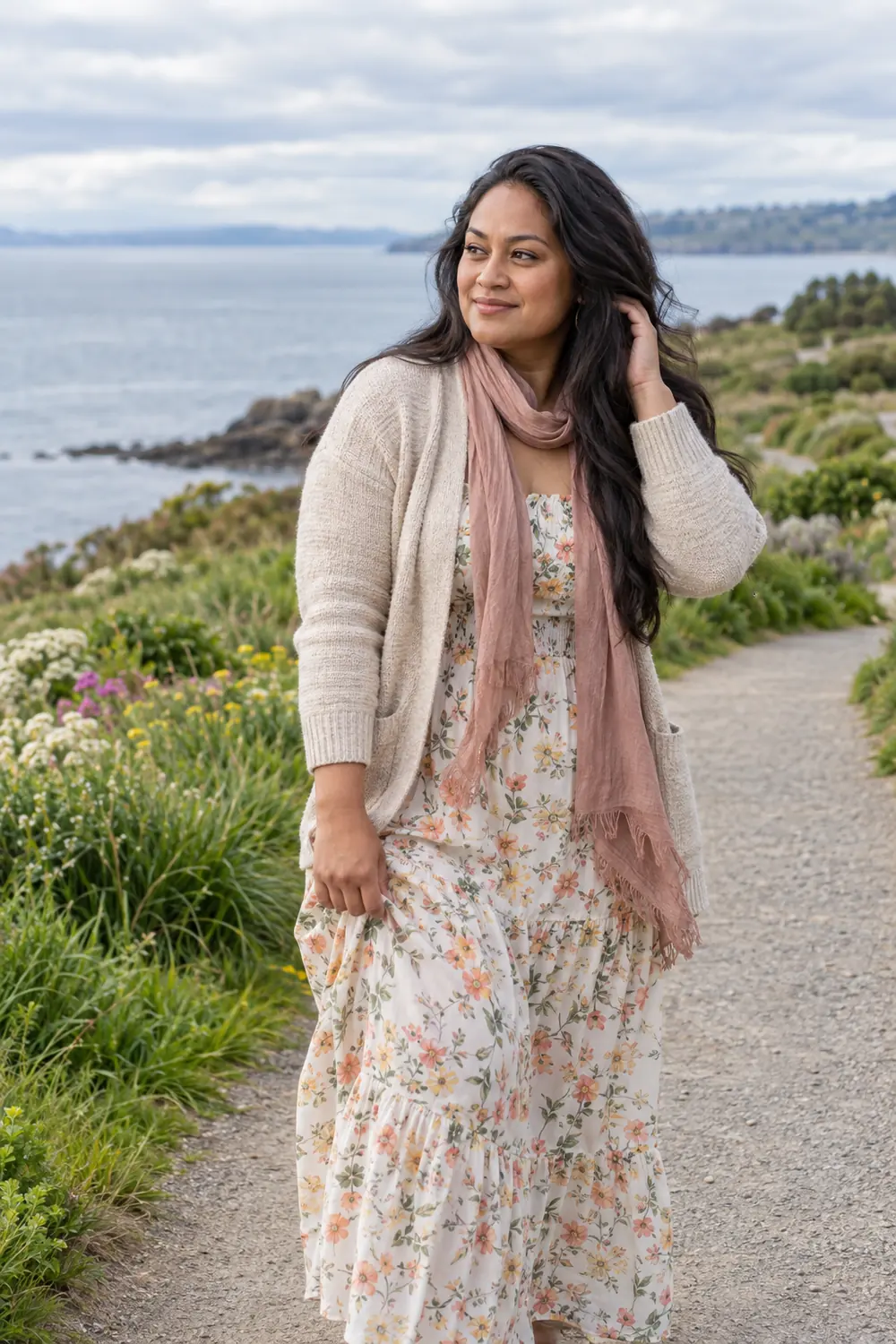 A woman with long dark hair stands on a wide coastal path in a floral dress, oatmeal cardigan and pink scarf, with spring flowers and calm water behind her.