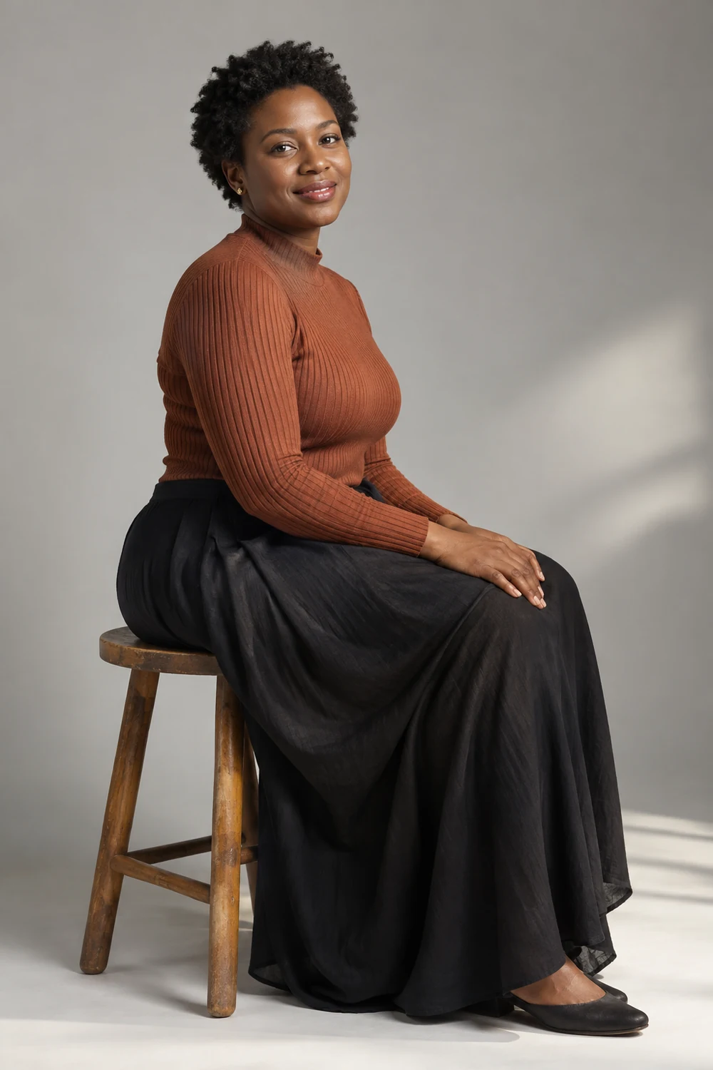 Adult woman with short coiled hair seated sideways on a wooden stool, wearing a rust knit top and long dark skirt against a softly lit gray studio backdrop.
