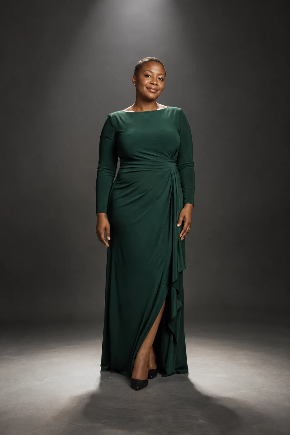 Adult woman with close-cropped hair in a deep green evening dress standing in a pool of light on a dark gray studio backdrop.