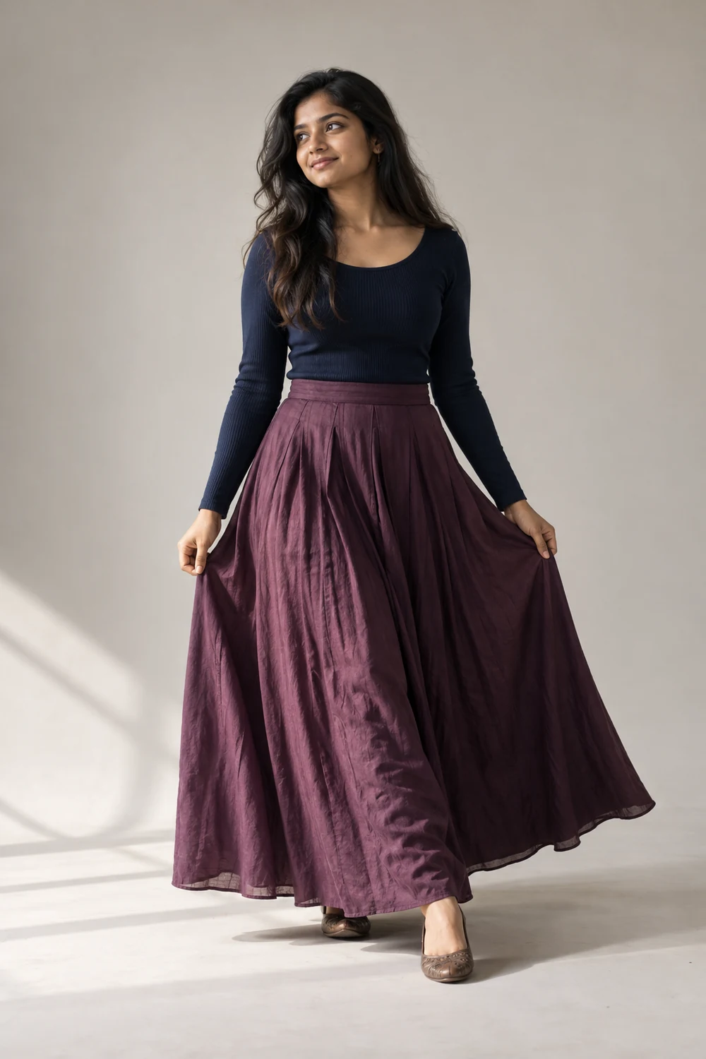 Adult South Asian woman in a navy long-sleeved top and flowing plum skirt, stepping and holding the skirt sides in a warm-gray studio.