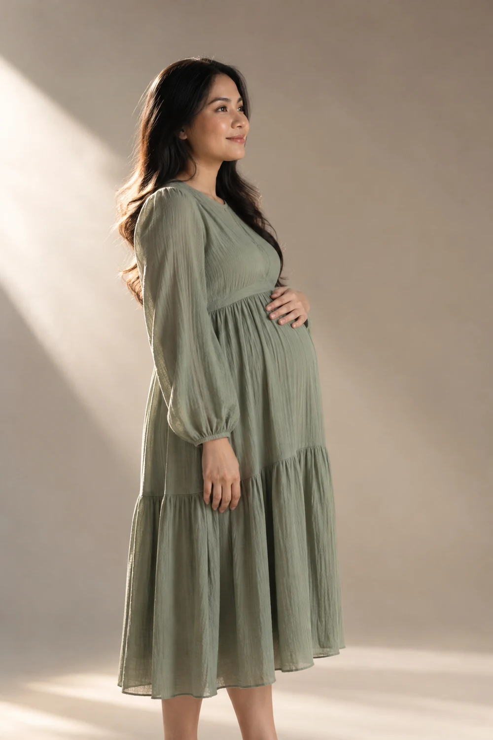 A visibly pregnant woman in a loose sage maternity dress stands in profile against a softly lit neutral studio backdrop.