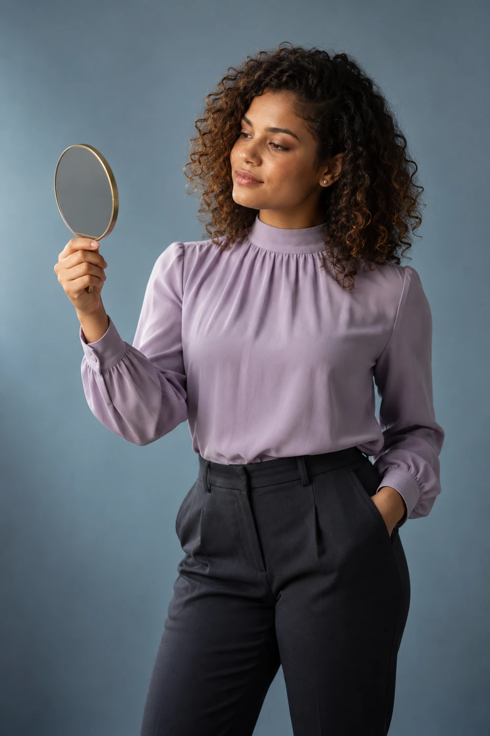 Woman with shoulder-length natural curls holding a round hand mirror at chest height in a blue-gray studio, wearing a lavender blouse and dark trousers.